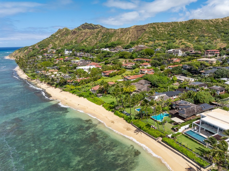 Aerial view of a beachfront neighborhood nestled between lush green mountains and turquoise ocean waters, showcasing the scenic coastal setting and surrounding natural landscape.