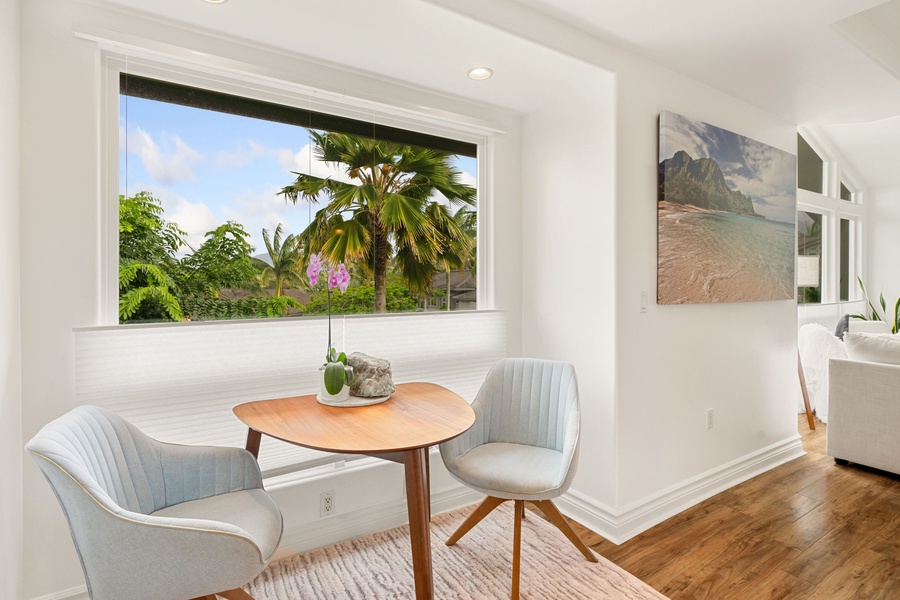 A serene corner of the primary suite boasts a minimalist wooden round table paired with a pastel blue upholstered chair, a perfect spot for your morning coffee w/ frames a picturesque view of lush palm trees.