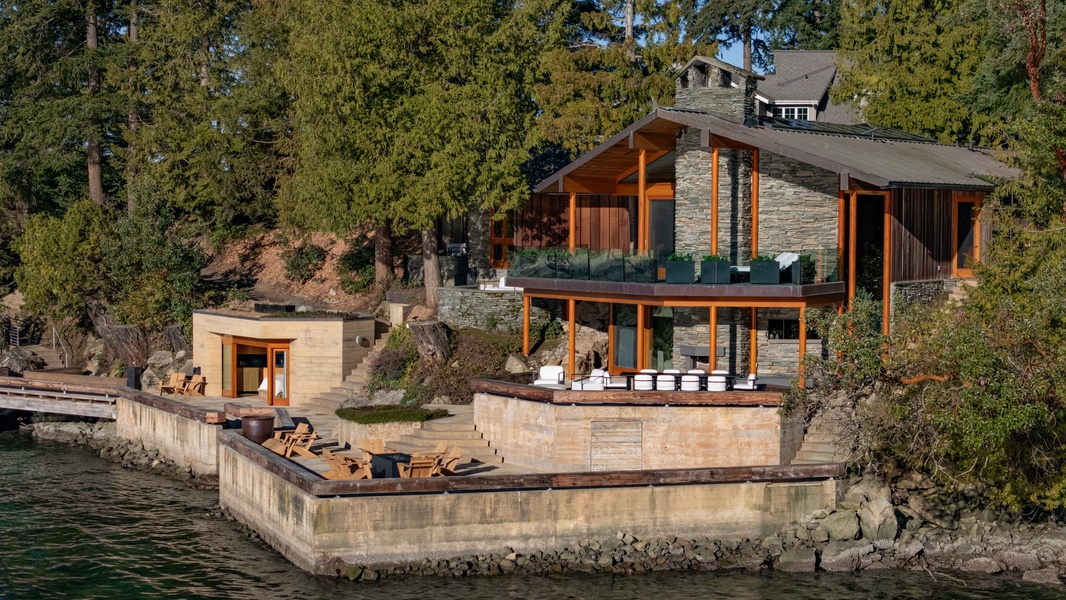 Modern lakefront retreat with stone and wood architecture, featuring expansive windows and waterfront deck surrounded by forest.