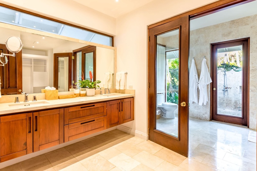 Modern ensuite full bath with a wooden double vanity and glass doors that lead to a lush outdoor shower area.