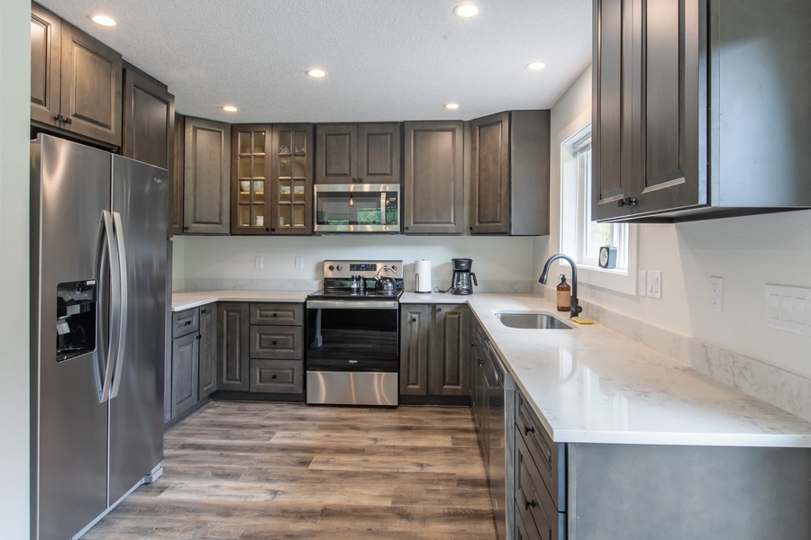 The fully equipped kitchen has all new stainless steel appliances, ample counter space, several culinary tools, a large island countertop with bar stool seating, and forest views
