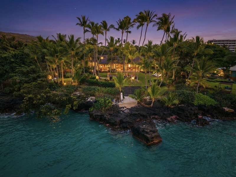 Hahalua Bay Villa sits oceanfront amid tropical palms with turquoise waters and volcanic rock formations creating a stunning Hawaiian setting.