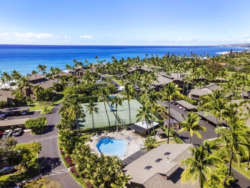 Stunning aerial view of a tropical resort complex featuring pristine blue ocean, palm-lined beach, swimming pool, and tennis court.