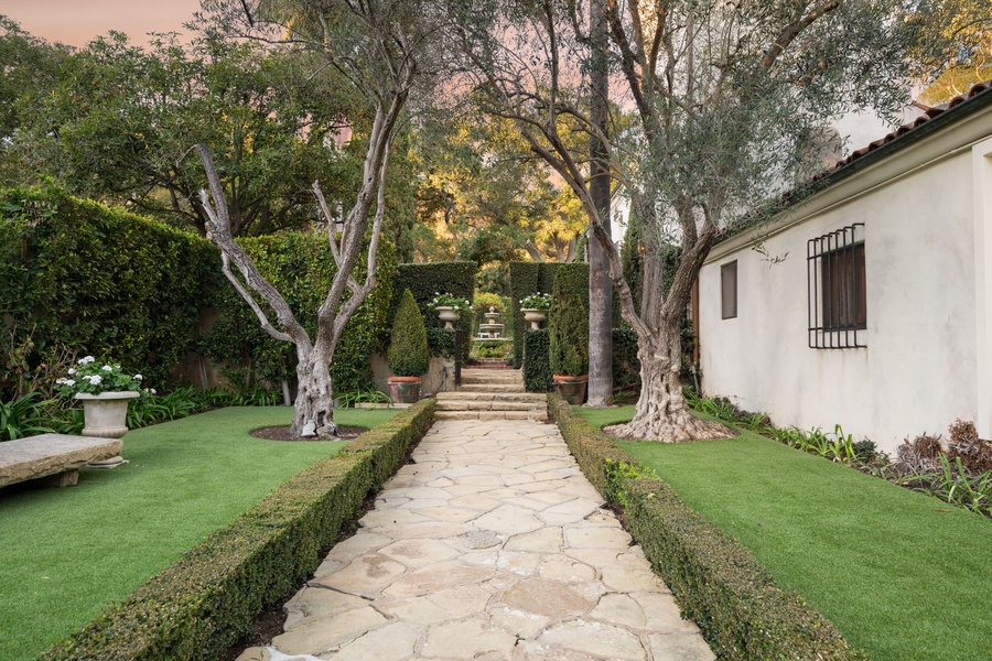 Manicured gardens with stone pathways lead through the property's elegant landscaped grounds, featuring mature trees and formal hedging.