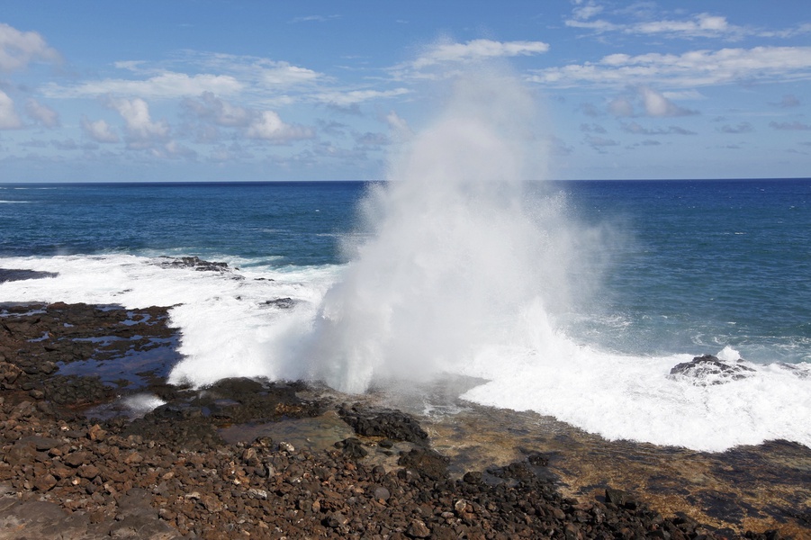 Dramatic ocean waves crash against rocky coastline, creating spectacular water geysers under bright blue skies.