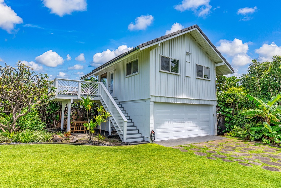 Elevated home with tropical landscaping, featuring a private garage and surrounded by lush greenery under clear blue skies.
