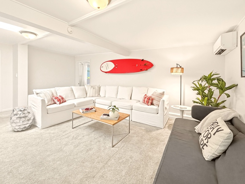 Sink into plush white sectional seating where surfboard art and tropical plants create your perfect coastal retreat atmosphere.