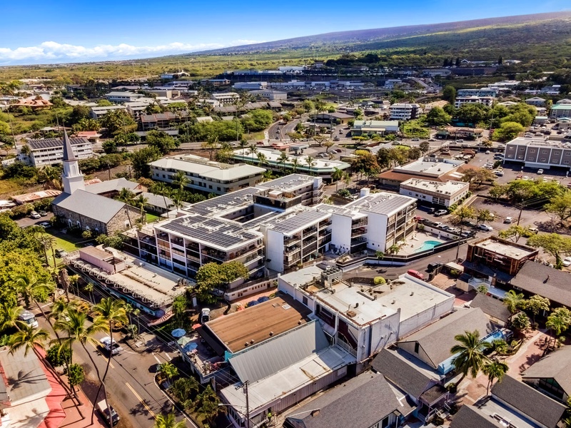 Aerial view of a charming tropical town with mix of modern buildings, local shops, and lush greenery beneath blue skies.