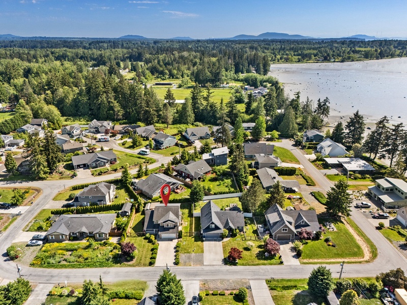 Aerial view of a residential neighborhood nestled along a scenic waterfront, surrounded by forests and distant mountain ranges under a clear blue sky.