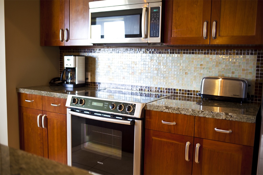 The kitchen is well-equipped with modern appliances, including a sleek stainless steel oven and microwave. The mosaic tile backsplash adds a touch of elegance, complementing the rich wood cabinetry and granite countertops.